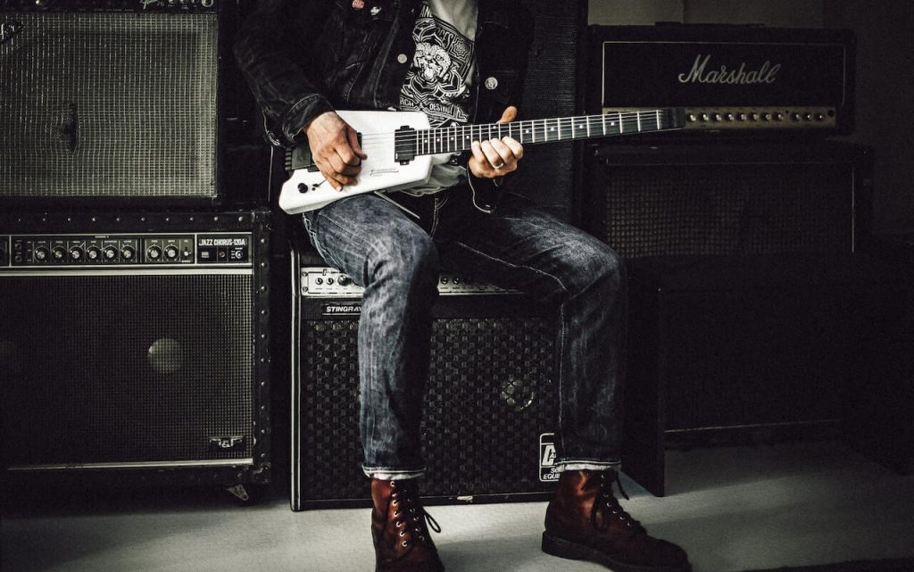 Un'immagine di un uomo seduto sopra un amplificatore per chitarra, che suona appassionatamente la chitarra.